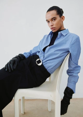 Person seated wearing a blue-striped shirt, black pants with a circular buckle, and long black gloves in a neutral indoor setting.