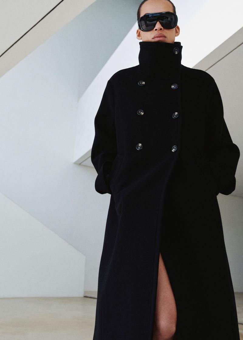 Fashionable adult woman wearing a black overcoat and sunglasses posing indoors against a bright white architectural backdrop.