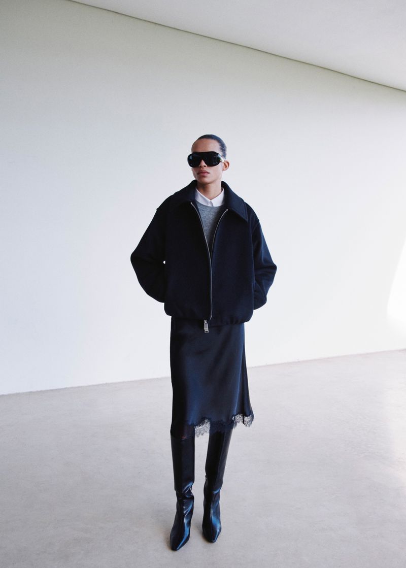 Model wearing black shield sunglasses, a boxy black zip jacket, black lace-trim midi skirt, and knee-high black leather boots.