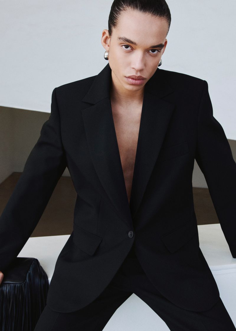 Adult androgynous fashion model poses in an elegant black suit, glancing at the camera in an indoor studio with neutral colors.