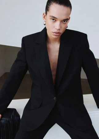 Adult androgynous fashion model poses in an elegant black suit, glancing at the camera in an indoor studio with neutral colors.