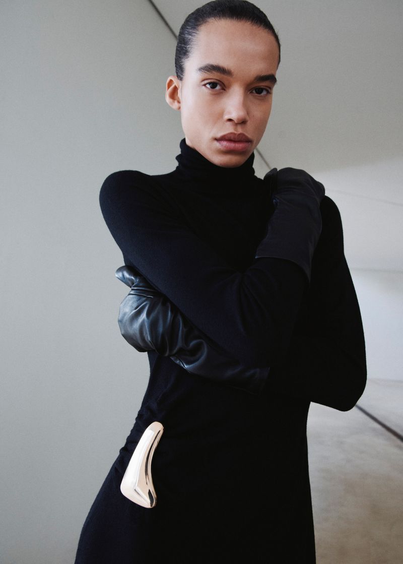 Fashion portrait of a slim adult woman with slicked-back hair in a black long-sleeve turtleneck dress and leather gloves indoors.