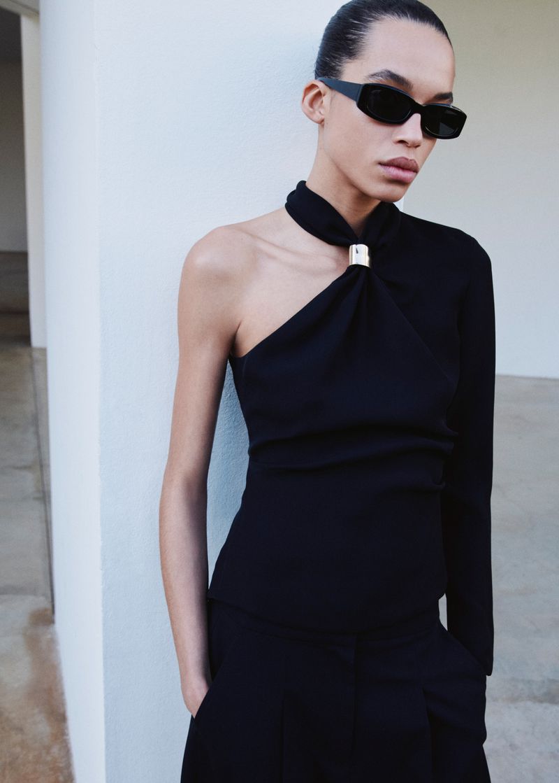 Woman in black one-shoulder top and pants poses against a light wall, wearing black sunglasses in a fashion shot.
