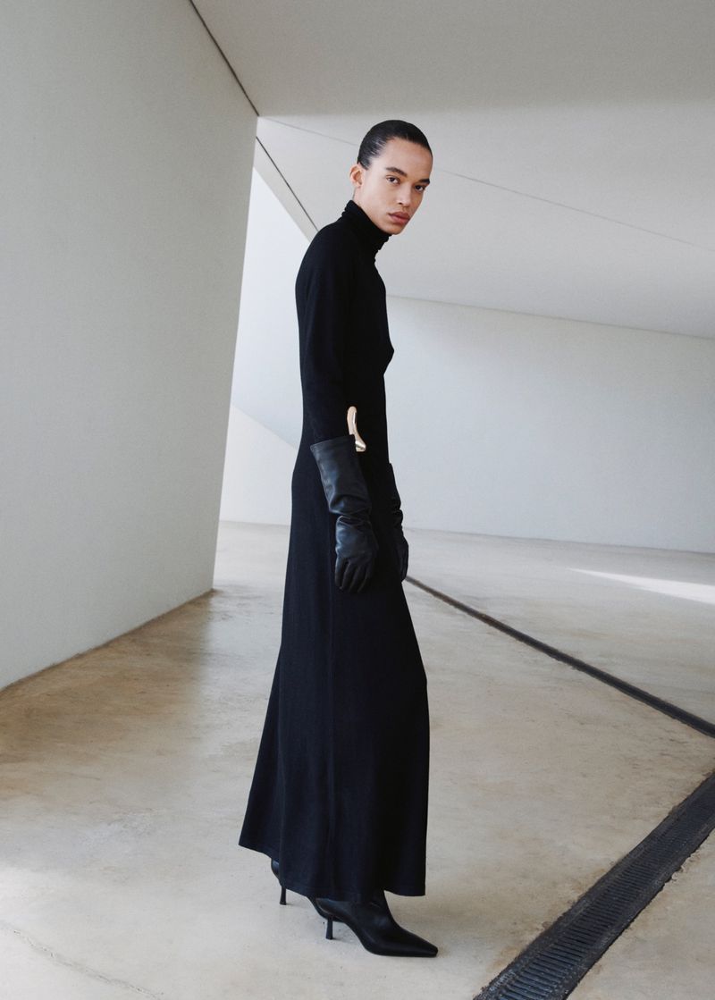 An adult female fashion model in a black turtleneck dress with gloves and boots, posing in an indoor minimal architectural space.