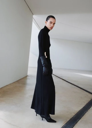 An adult female fashion model in a black turtleneck dress with gloves and boots, posing in an indoor minimal architectural space.