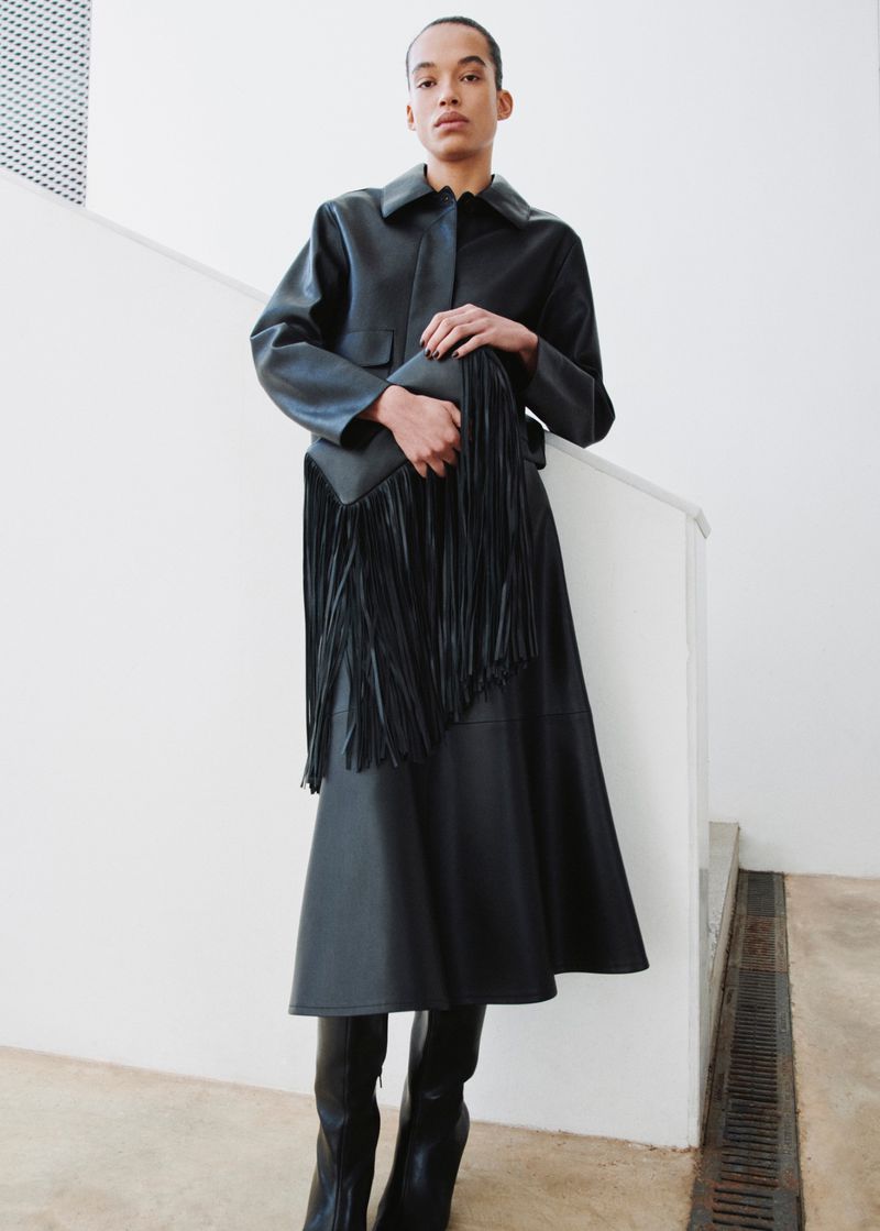 An adult female posing indoors wearing a black leather-like jacket, A-line skirt, high heeled boots, and holding a fringed purse.