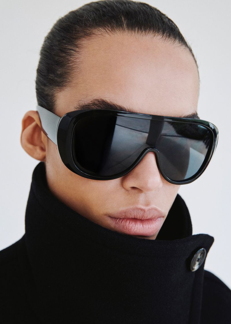 Close-up portrait of a model wearing black sunglasses and a dark coat against a plain white background.