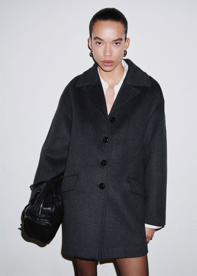 Woman in a dark grey wool coat and black tights standing against a white wall, holding a large black leather handbag.