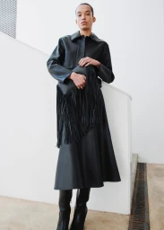 Model standing on a staircase wearing a black leather jacket, midi skirt, and boots while holding a fringed clutch.