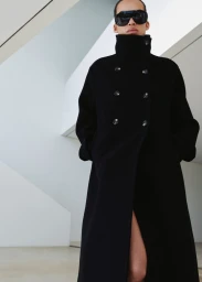 Model wearing a long black double-breasted coat with a high collar and black shield sunglasses against a white geometric interior.