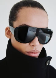 Close-up portrait of a model wearing black sunglasses and a dark coat against a plain white background.