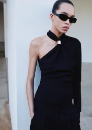 Model wearing dark sunglasses and an asymmetric one-shoulder black top leaning against a white architectural pillar.