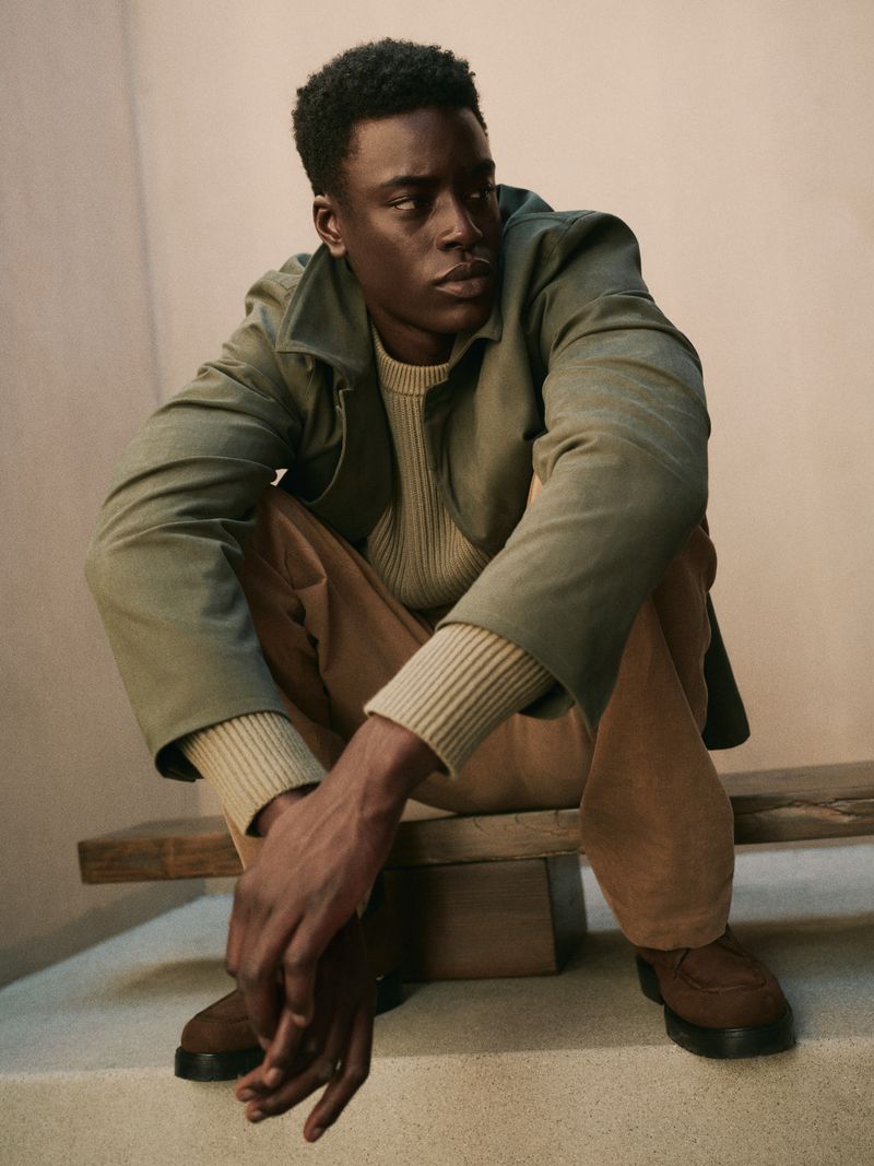 An African American man sits on a rustic bench indoors, wearing a green jacket, cream sweater, brown pants, and matching dress shoes in a portrait.