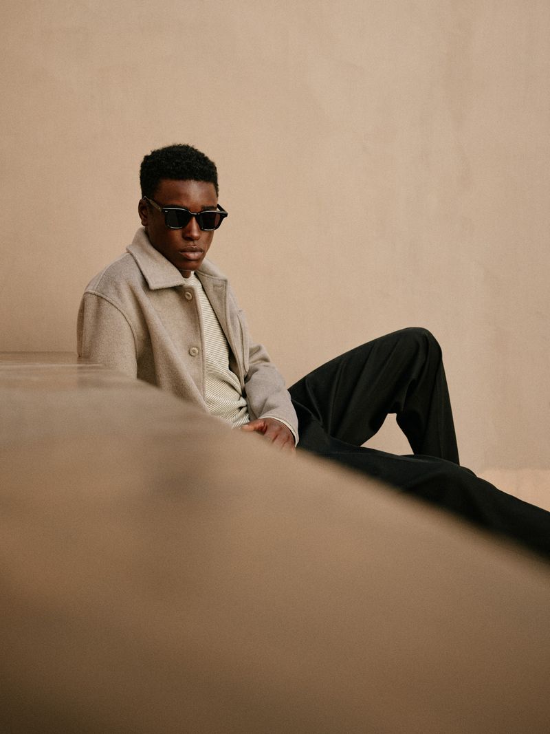 Man in sunglasses and beige jacket poses indoors. Neutral tones and a minimalist aesthetic create a sophisticated fashion image.