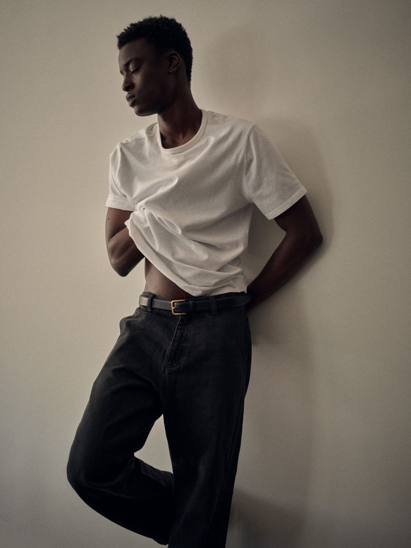 Adult male model with dark hair poses leaning against a wall. He wears a white t-shirt and dark-wash jeans with a gold belt buckle.