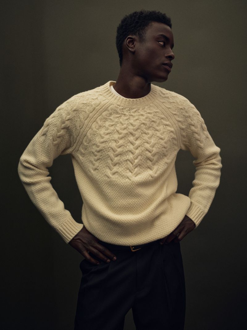 Babacar N'doye wearing a cream textured knit sweater and dark trousers against a muted dark studio background.