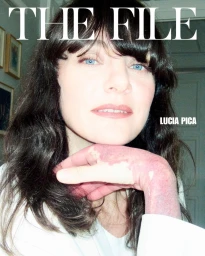 Close-up portrait of a woman with dark wavy hair and blue eyes resting her chin on her hand, featuring the text The File and Lucia Pica.