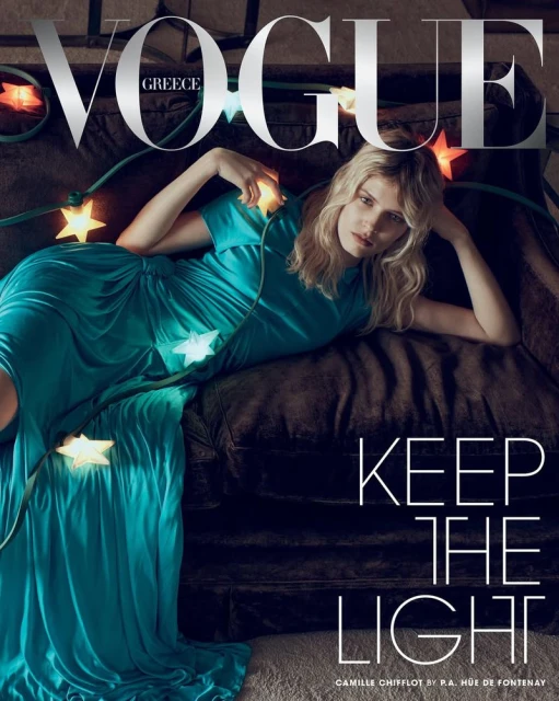 Model Camille Chifflot in a draped teal satin gown reclining on a dark couch adorned with glowing star string lights for Vogue Greece.