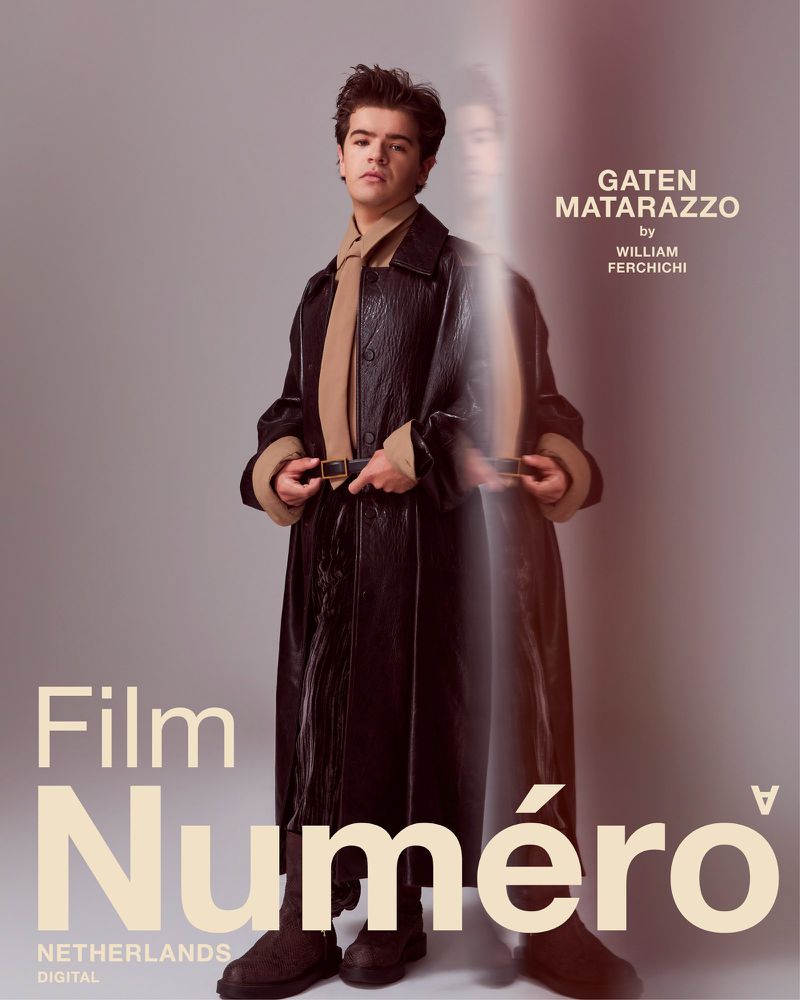 Portrait of Gaten Matarazzo wearing a dark brown, long leather coat with a tan scarf and dark brown boots against a grey background.