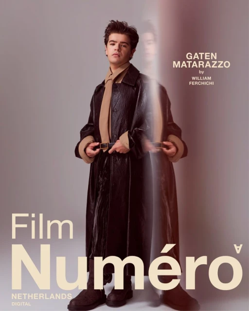 Portrait of Gaten Matarazzo wearing a dark brown, long leather coat with a tan scarf and dark brown boots against a grey background.