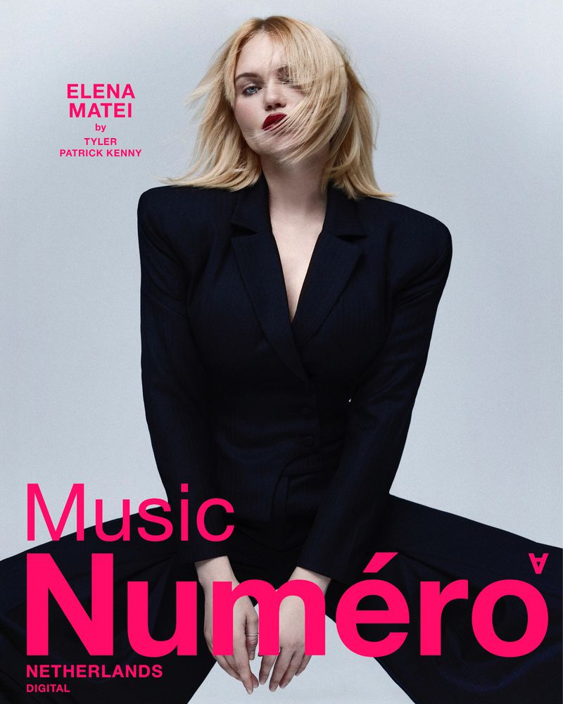 A woman poses in a dark blazer on a light background with pink magazine text overlaying her lower body in a studio shot.