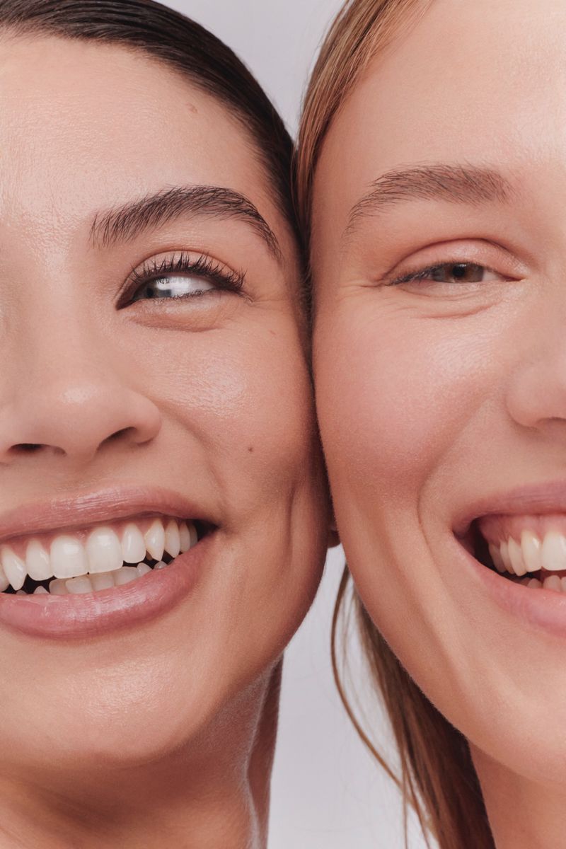 Close-up portrait of two women, one with dark hair and the other with blonde hair, smiling against a plain light gray studio backdrop.