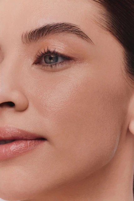 Close-up of a woman's face with clear skin and natural makeup. Focus on her blue eye, eyebrow, cheek, and lips against a neutral background.