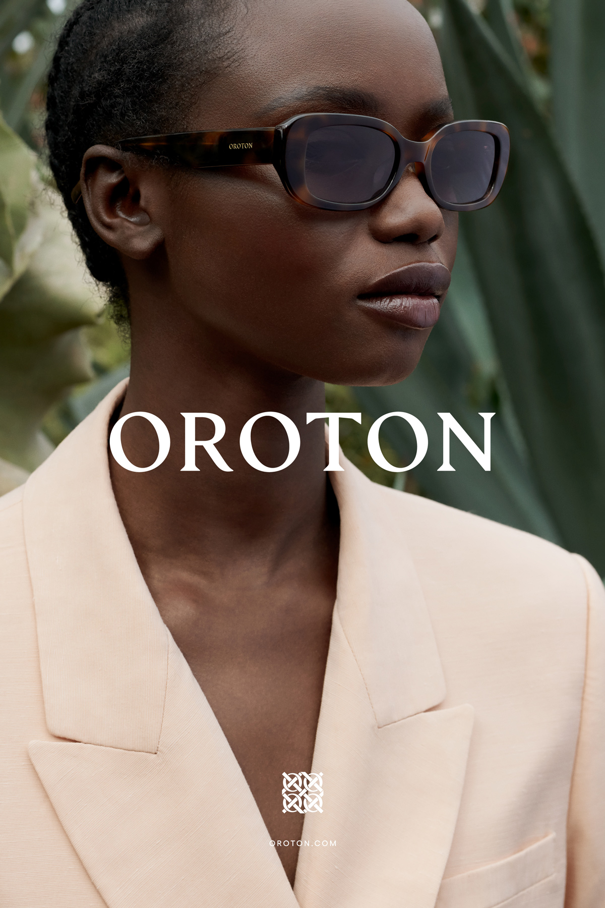 A dark-skinned woman with sunglasses models a beige blazer outdoors with palm fronds in the background for Oroton advertising campaign.