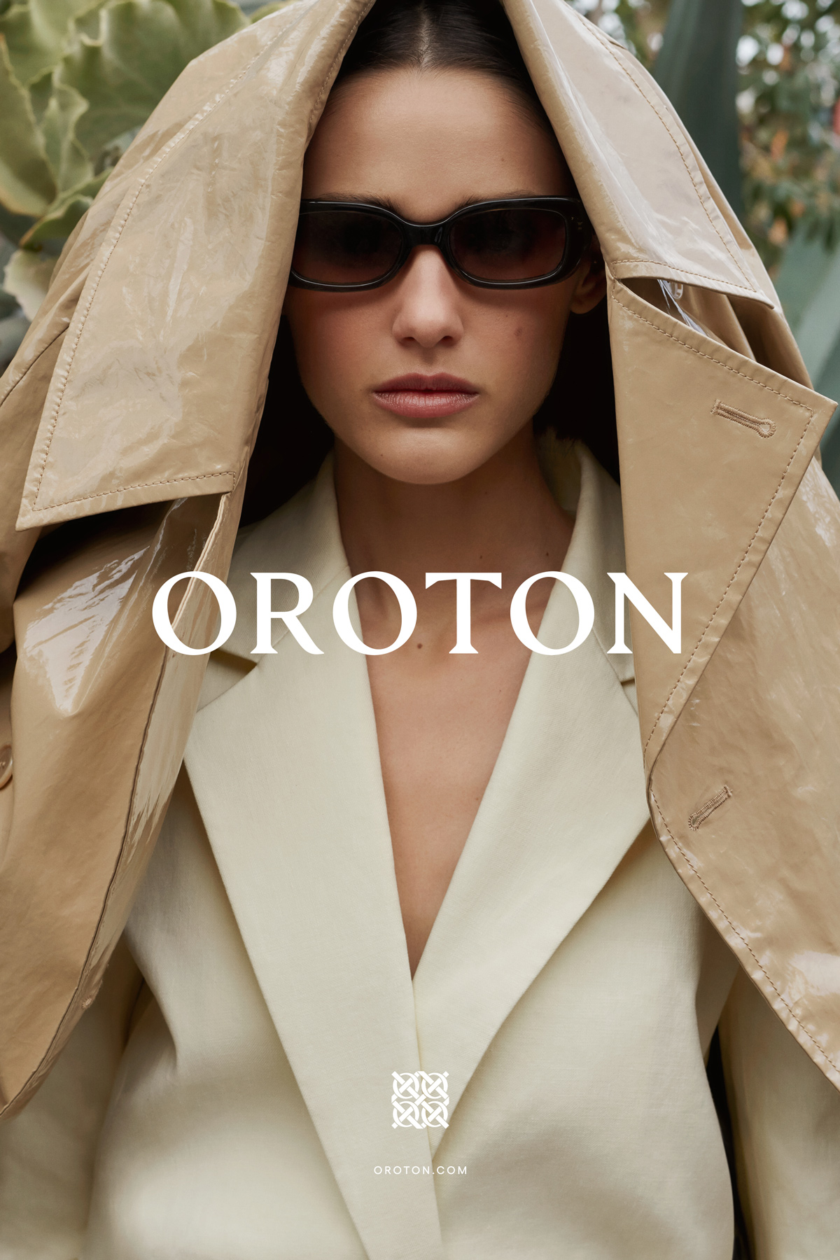 Fashion shot of a woman wearing sunglasses, an open blazer, and a coat on her head, posing in front of leafy plants.