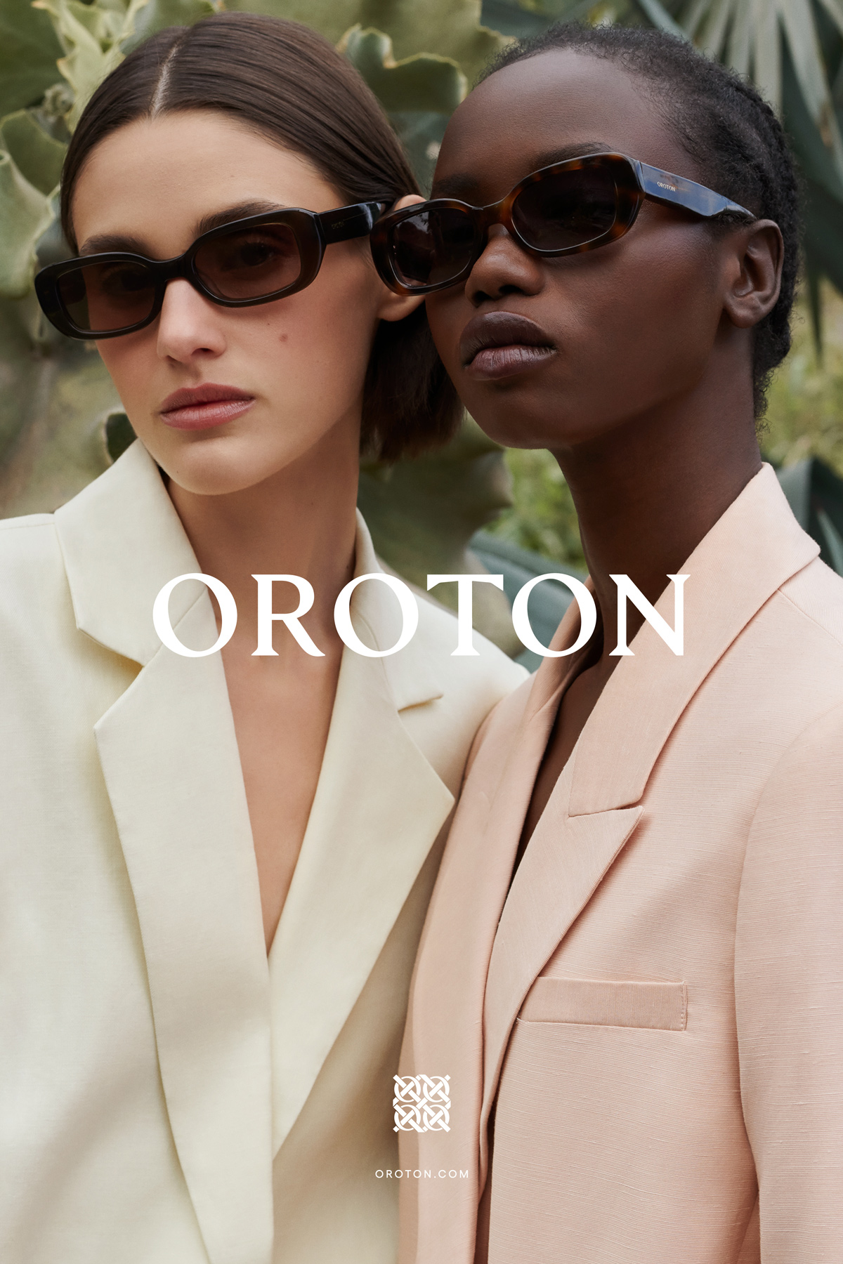 Two fashion models wearing sunglasses and blazers pose outdoors. The image showcases a sophisticated and trendy style with a natural backdrop.