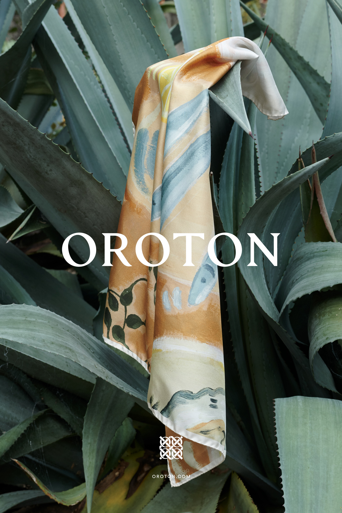 An abstract-print scarf from Oroton drapes among spiky green plants outdoors, creating a soft fashion accent against nature.