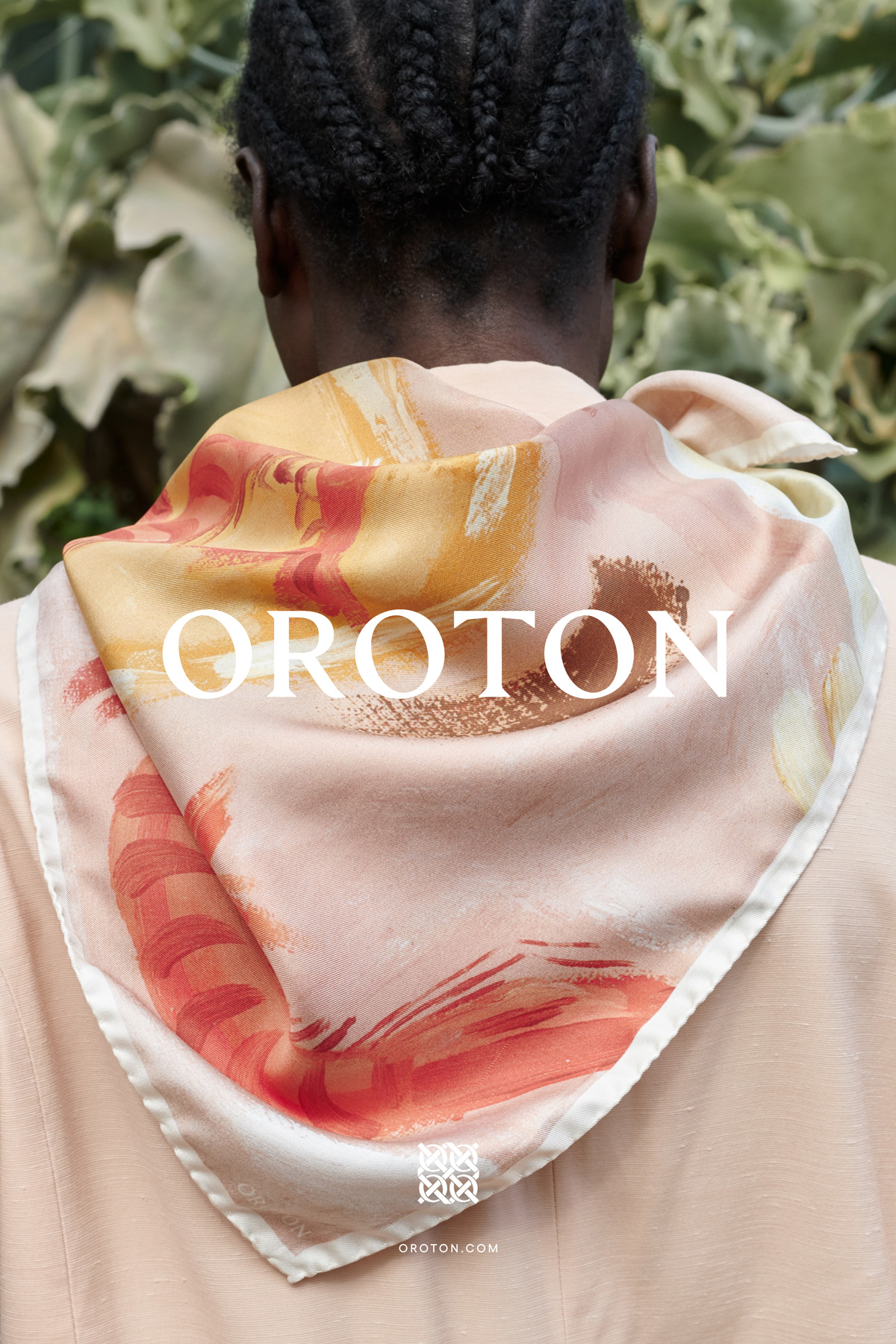 Back view of a person with braided hair wearing a patterned silk scarf, draped over their shoulders. Leafy green background.