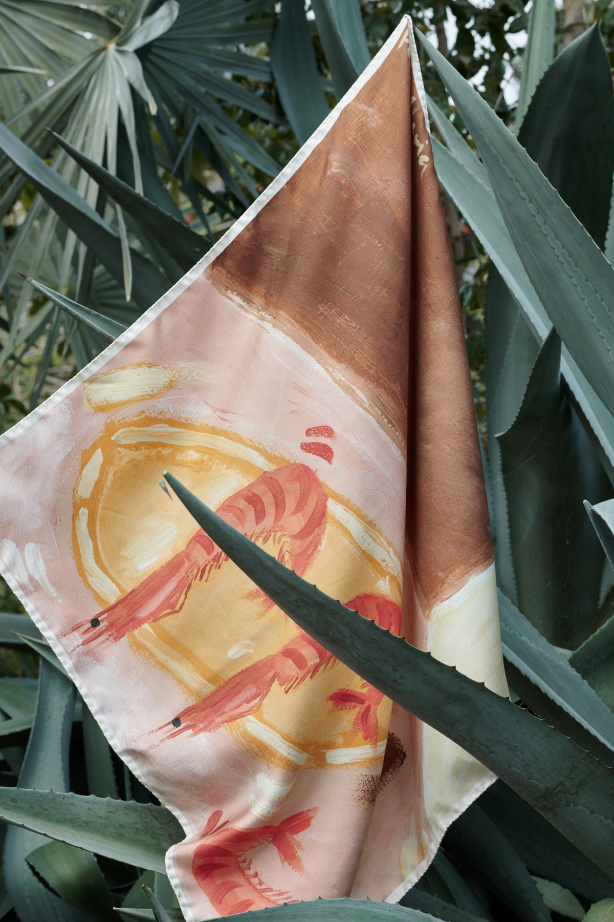Scarf featuring a shrimp illustration, draped over an agave plant, surrounded by lush green foliage. The background is vibrant green.