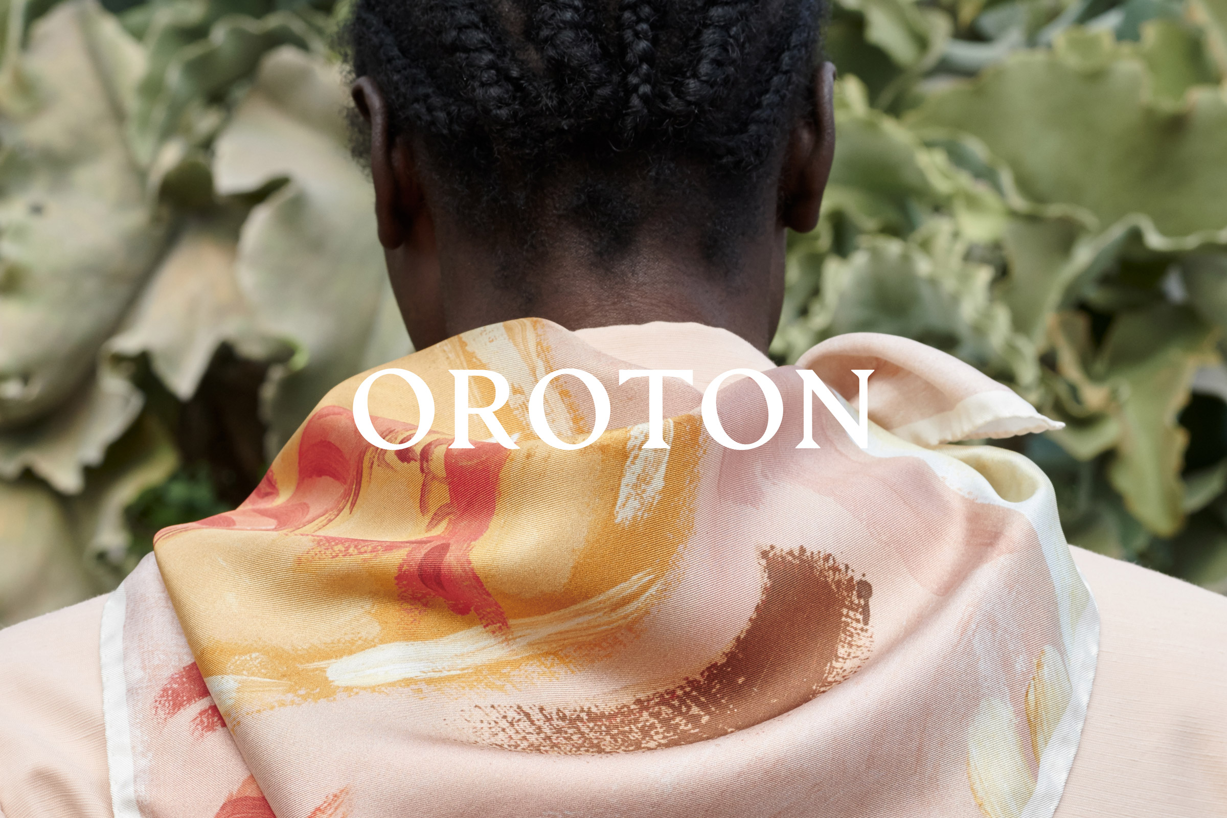 A person from the back wearing a patterned scarf with 'OROTON' text against a natural green background, focused on fashion and style.
