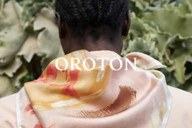 Rear view of a person wearing a patterned silk scarf with orange and cream brushstroke designs against a natural green foliage background.