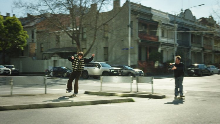 Young skateboarder in striped jacket performs trick on city street, filmed by another man with camera, urban setting on sunny day.