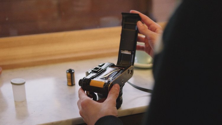 Person loading film into a vintage camera indoors. Visible are the camera body, hands, film cartridge, and canister on a light-colored surface.