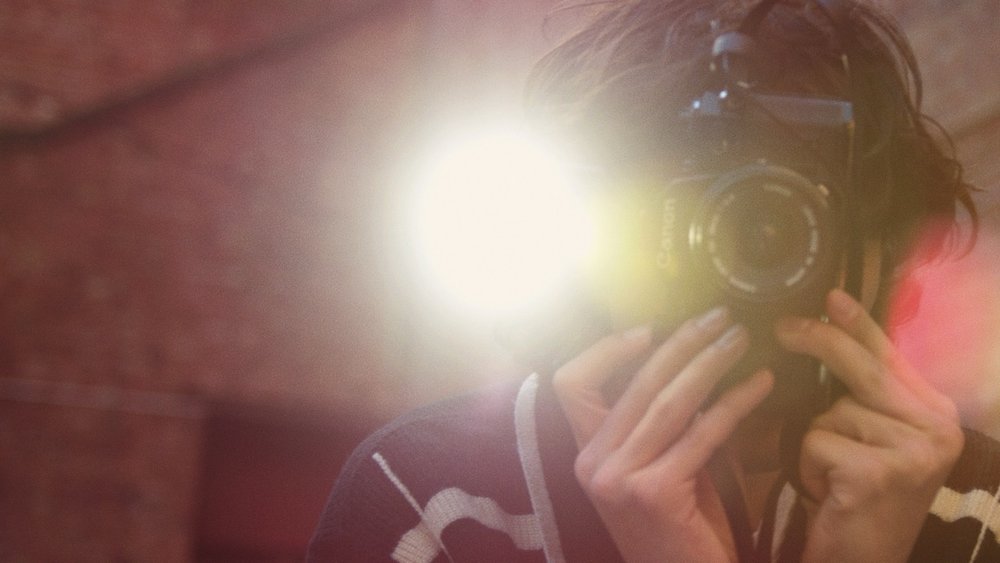 Adult person takes a photograph of themselves using a digital camera with a flash. The background is out of focus and reddish.