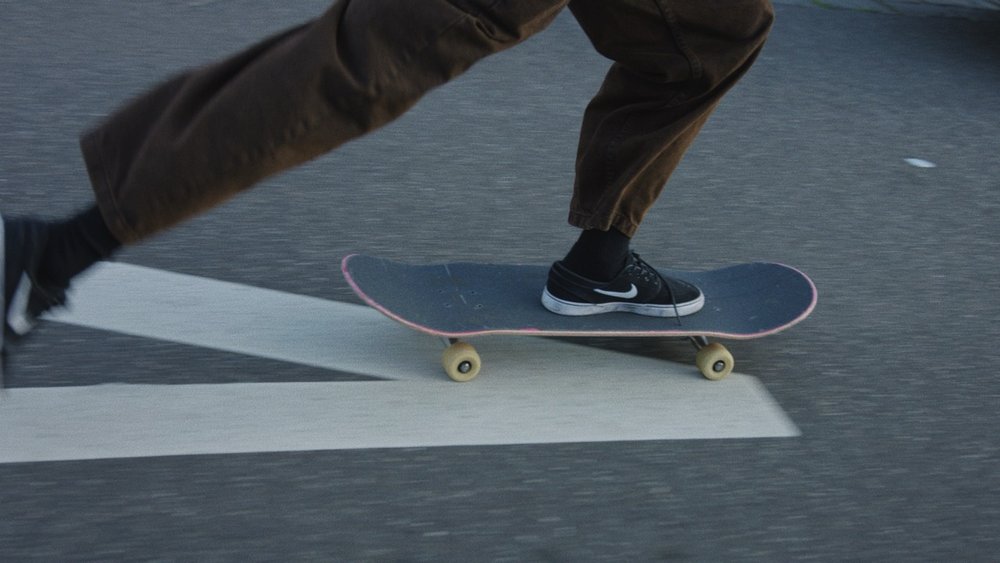A skateboarder rides on grey asphalt, captured at a medium angle showing brown pants, black sneakers, and the skateboard's motion.