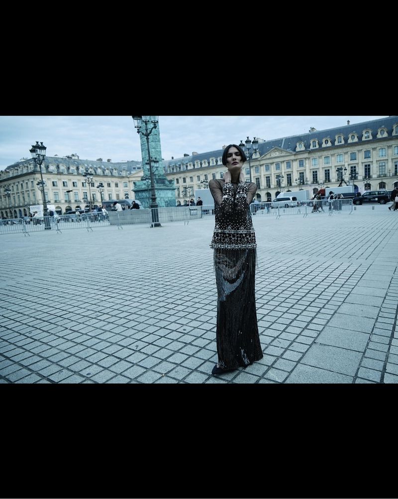 Woman in a gown poses in a city square with historic buildings in the background. The cobblestone square is empty under a blue sky.