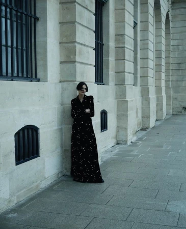 A stylish woman in a long black dress stands against a stone wall with barred windows, casting shadows on a stone-tiled ground.