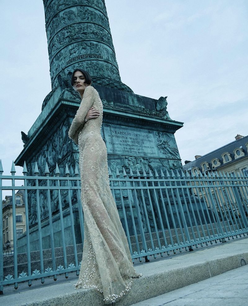 A woman in a formal beaded dress poses near a monument with an ornate iron fence. The Parisian architecture appears in the background.