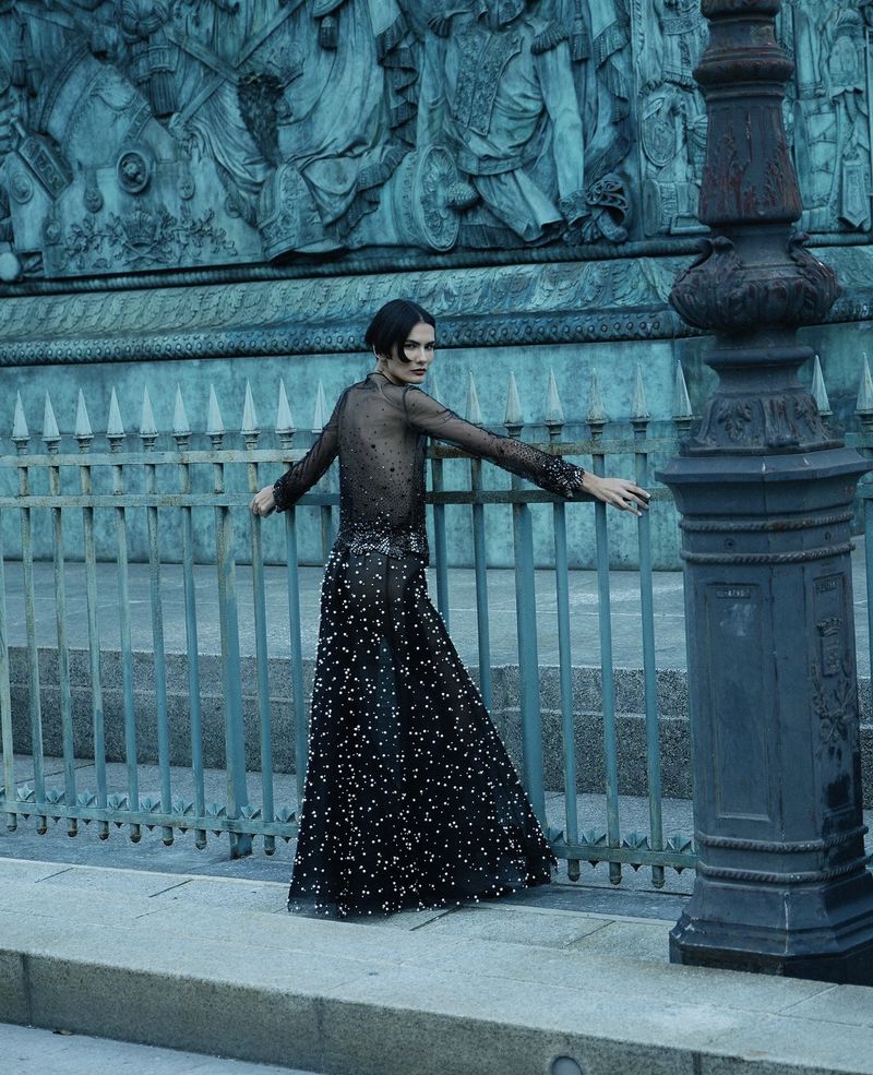 Fashion shoot of a brunette woman in a black, beaded gown, leaning against a rail with a building facade in the background.