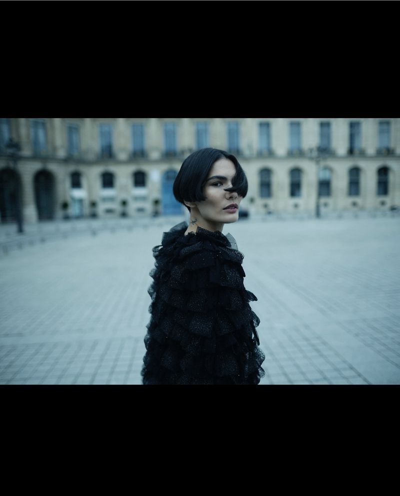 A woman with short dark hair and a black ruffled shawl stands in a plaza in front of a building with arched windows.