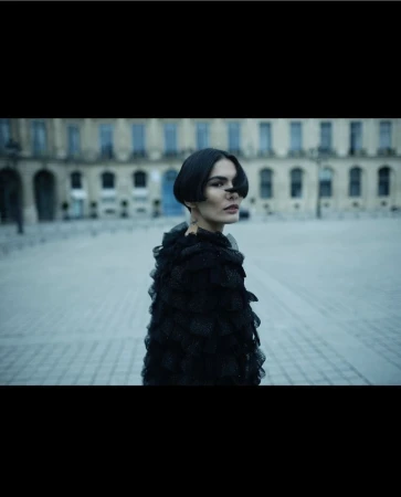 A woman with short dark hair and a black ruffled shawl stands in a plaza in front of a building with arched windows.