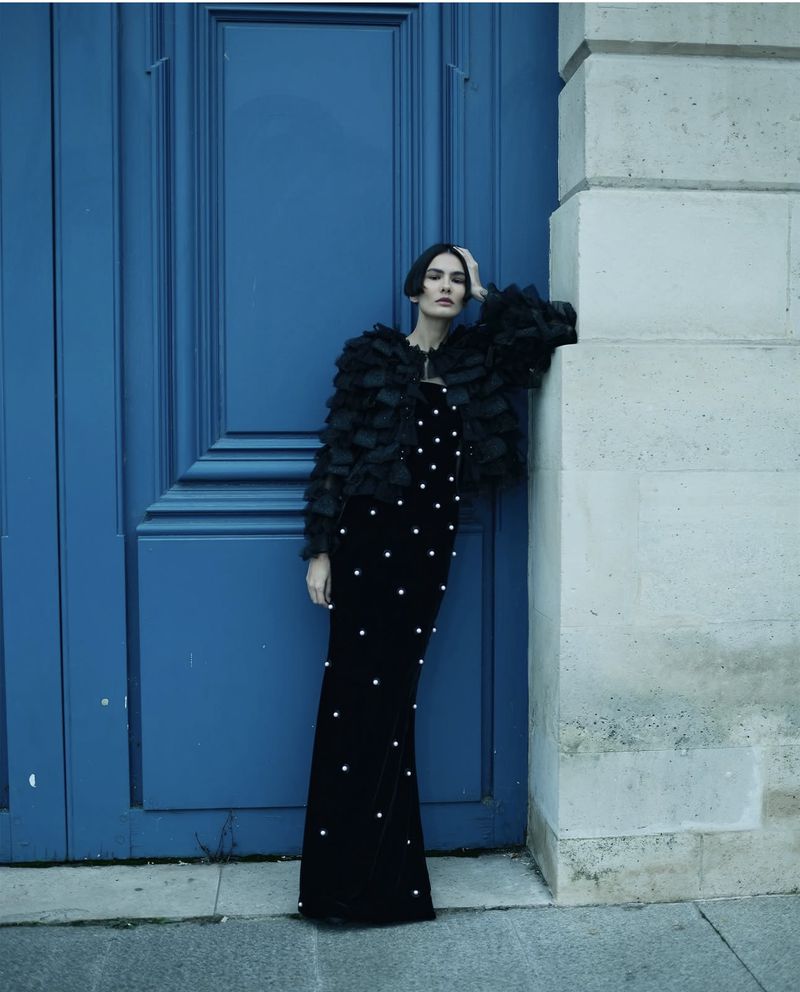 A woman poses against a blue door and a light-grey stone wall, wearing a black gown and jacket with pearl and ruffle details.