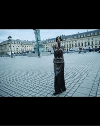 Model wearing dark embellished evening gown standing in a large cobblestone public square with classic architecture.