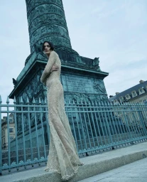 Model wearing a beaded long-sleeved gown standing outdoors before a towering bronze column and metal fence for Numéro Brasil.