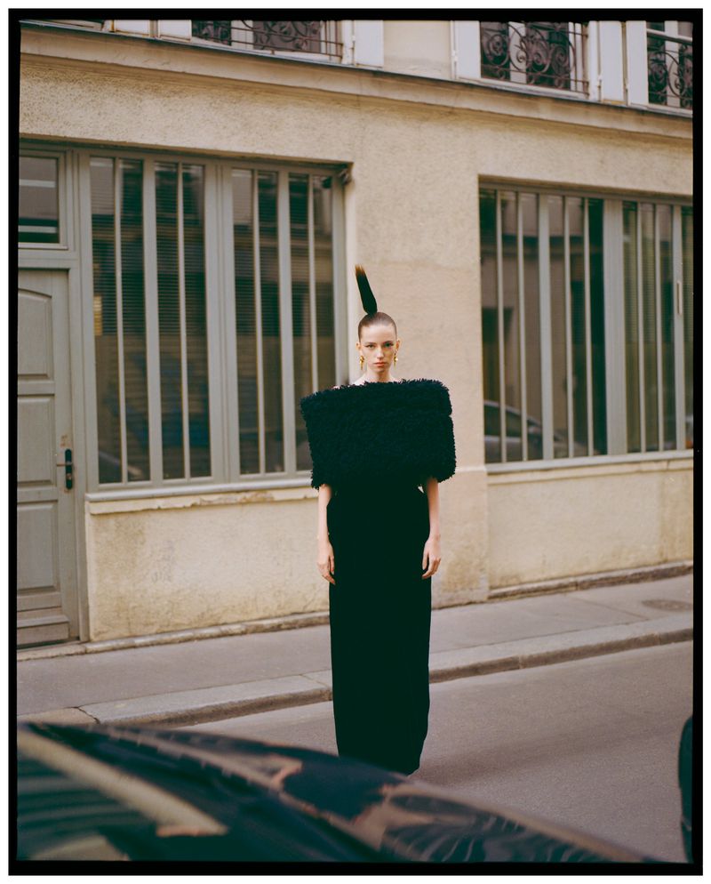 Fashionable woman in a black gown with sculpted shoulder detail poses on a city street in front of an apartment building. Editorial fashion.