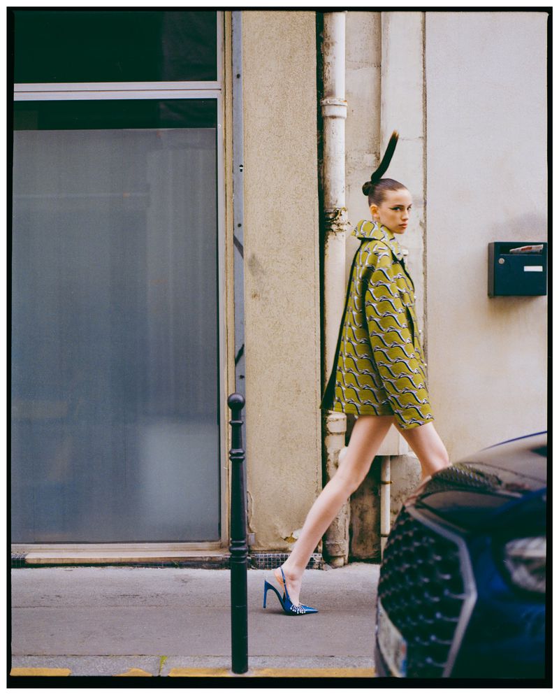 An adult female model walks on a sidewalk next to a building wearing a patterned green jacket and blue heels, with a dark blue car visible.